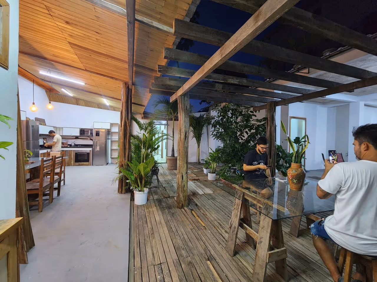 Área externa com decking de madeira, plantas em vasos e homens usando celulares sentados em banco e cadeira.