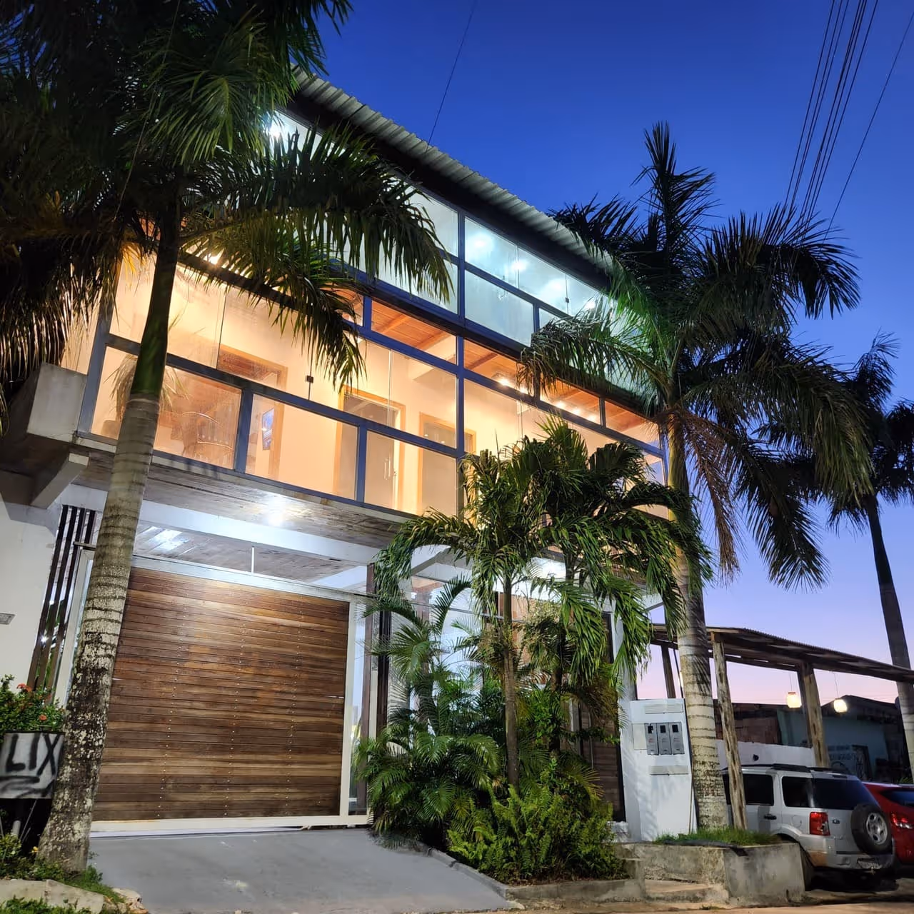 Fachada de casa moderna com grandes janelas e varanda iluminada, cercada por palmeiras.