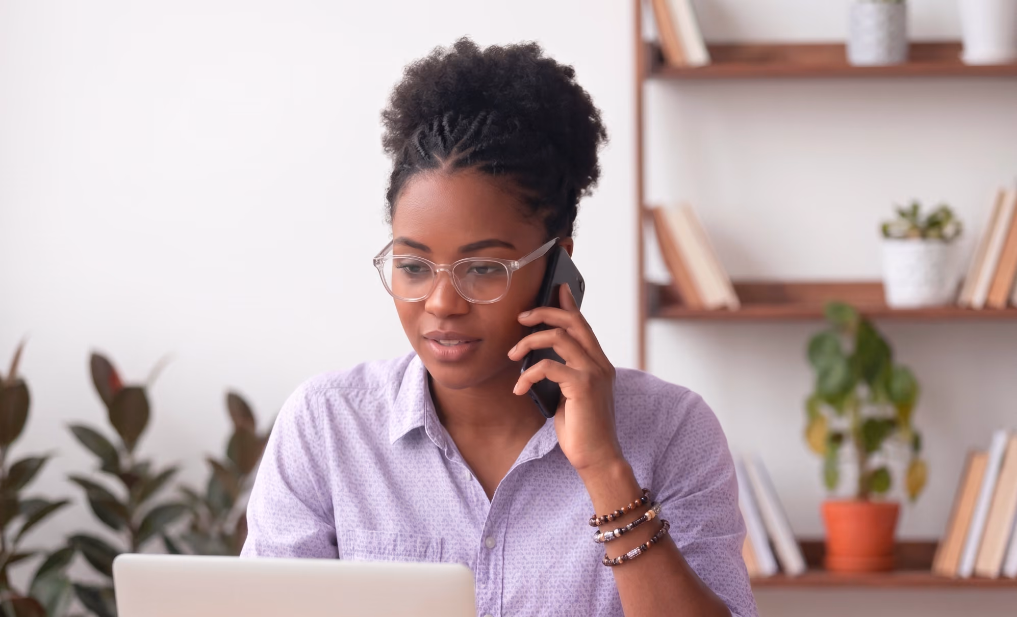 Mulher negra com óculos falando ao celular enquanto trabalha no laptop em ambiente com plantas e prateleiras.