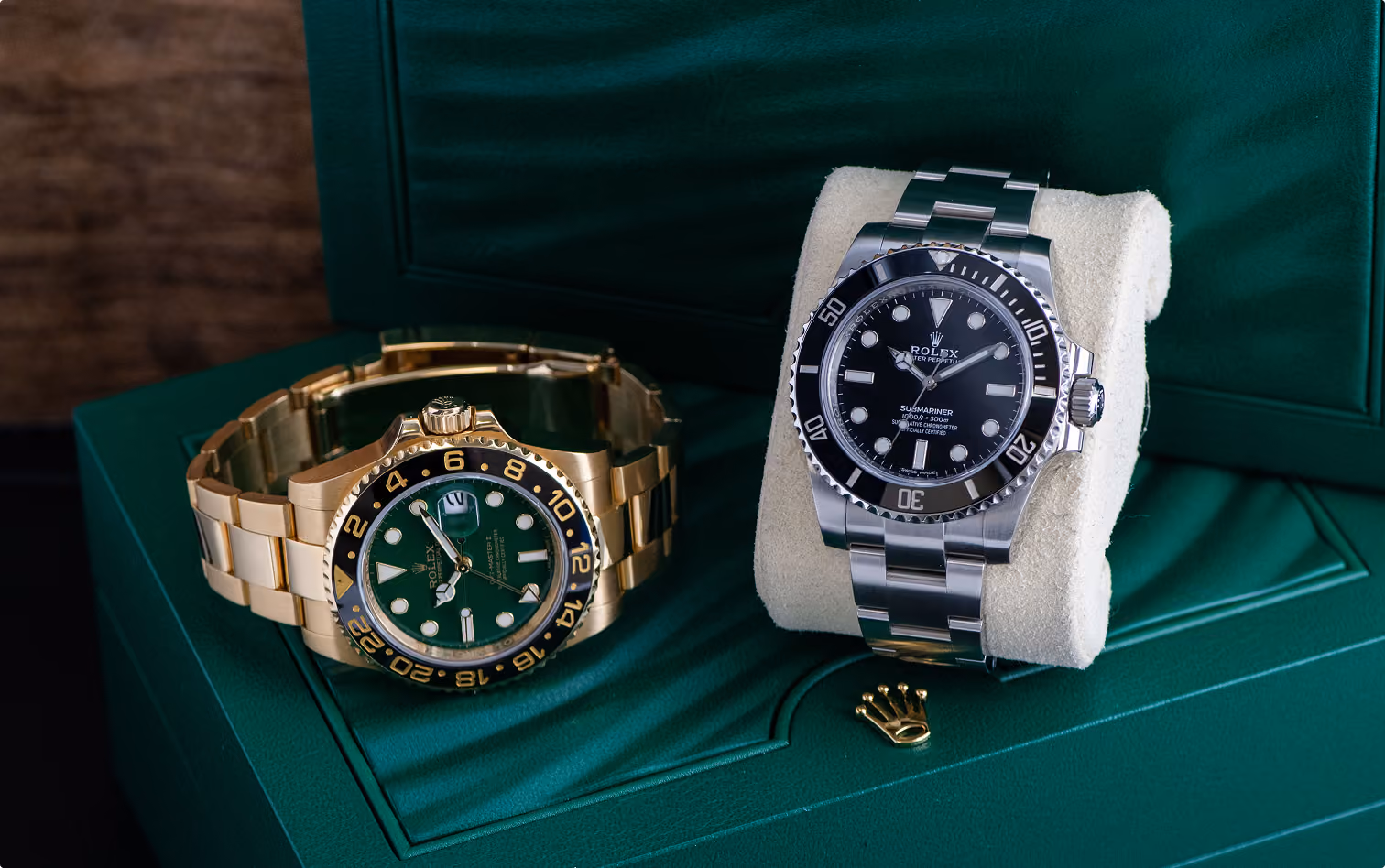 Two Rolex wristwatches displayed on a green leather box: one gold with a green dial and black bezel, the other stainless steel with a black dial and bezel on a beige cushion.