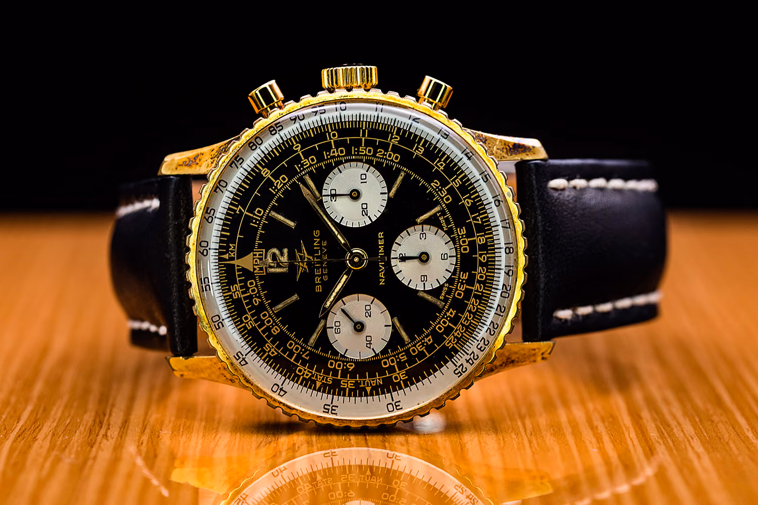 Close-up of a Breitling Navitimer watch with gold bezel and black leather strap on wood table.
