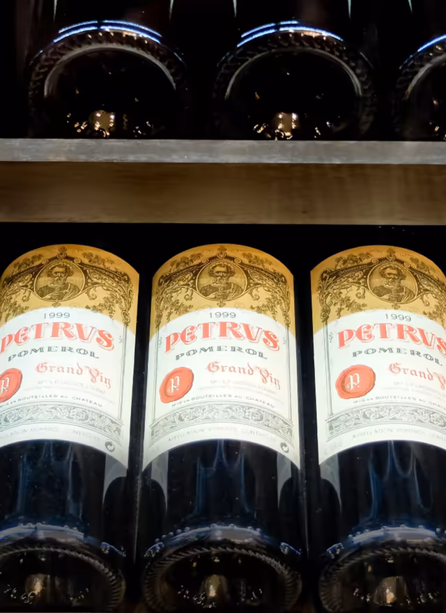 Three bottles of 1999 Petrus Pomerol Grand Vin wine with detailed gold and white labels, arranged side by side on a shelf with other bottles above.