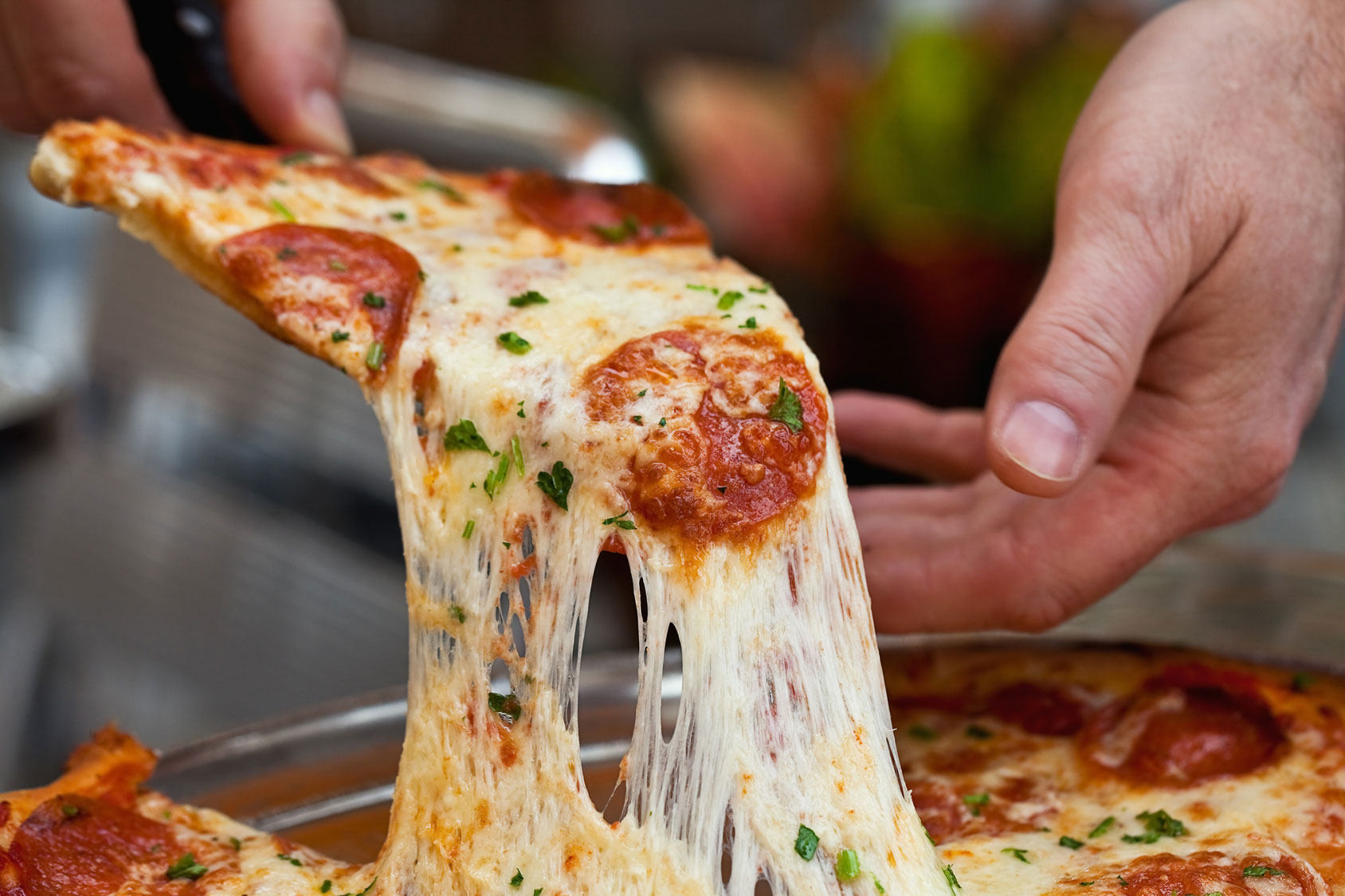 Hand lifting a slice of pepperoni pizza with melted, stretchy cheese and sprinkled parsley.