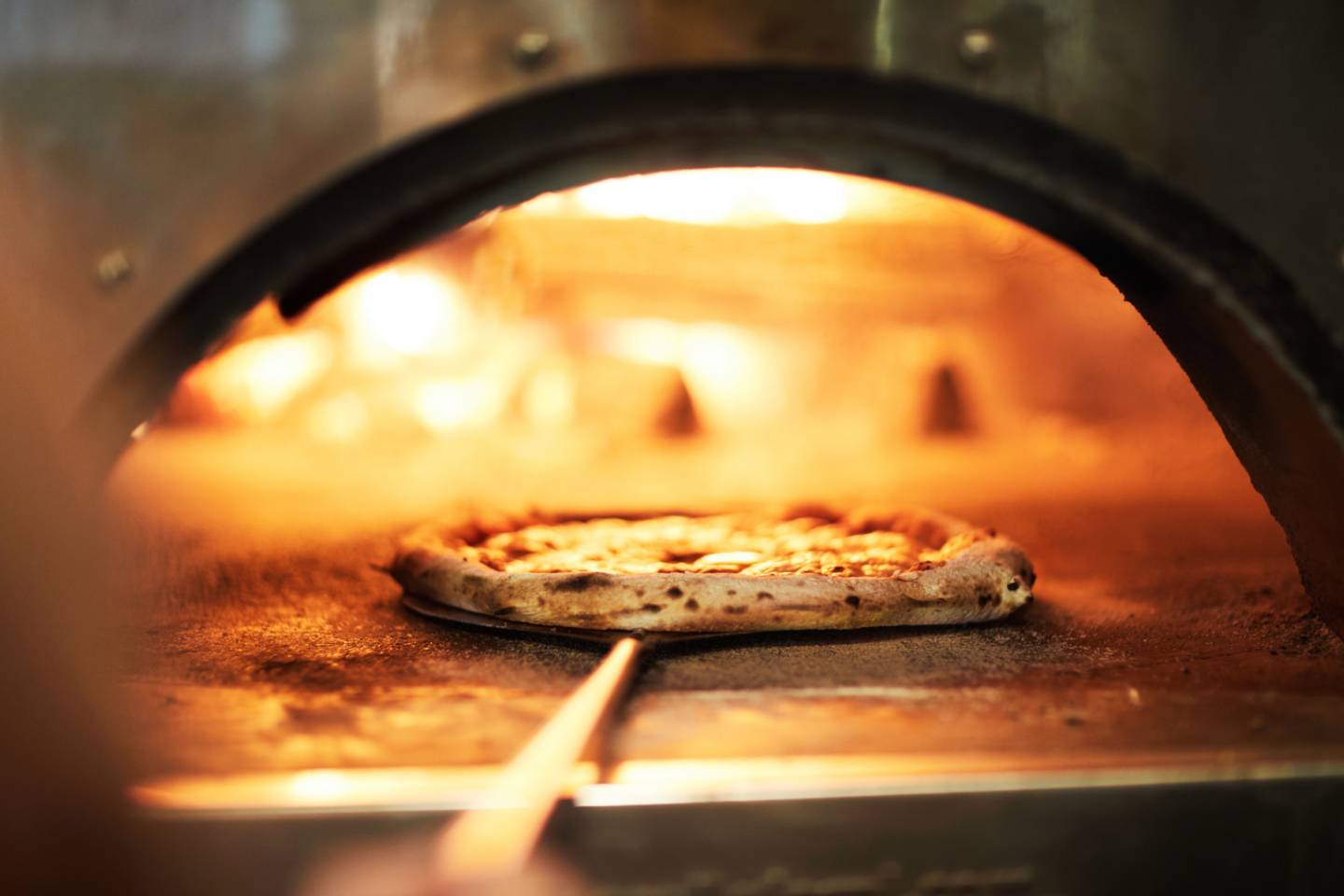 Pizza baking inside a wood-fired oven on a pizza peel.