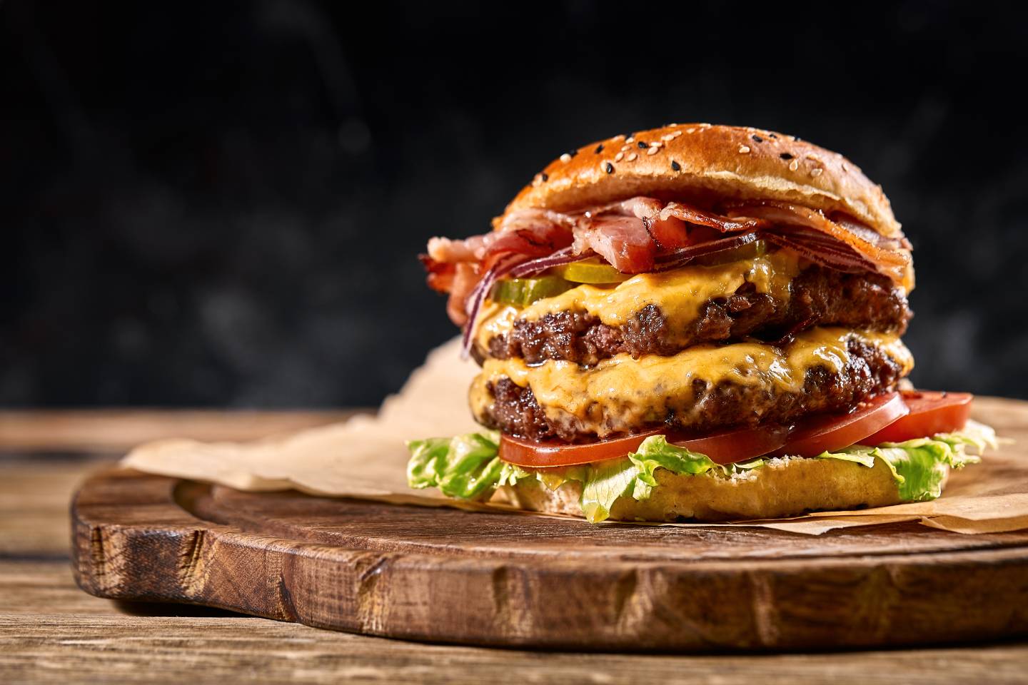 Double cheeseburger with bacon, pickles, tomato, lettuce, and sesame seed bun on a wooden board.