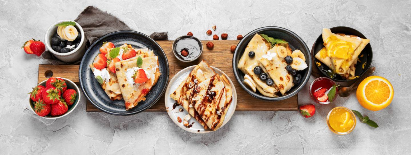 Plates of crepes topped with strawberries, blueberries, bananas, and orange slices, accompanied by bowls of fresh strawberries and blueberries, small glasses of orange and strawberry juice, and a half orange on a gray surface.