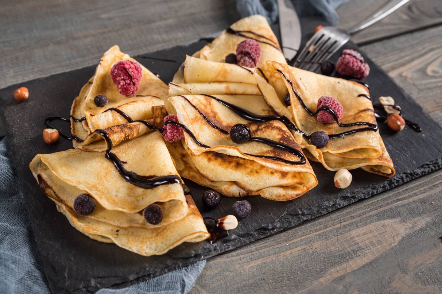 Folded crepes drizzled with chocolate sauce and topped with frozen raspberries and blueberries on a dark slate serving board.