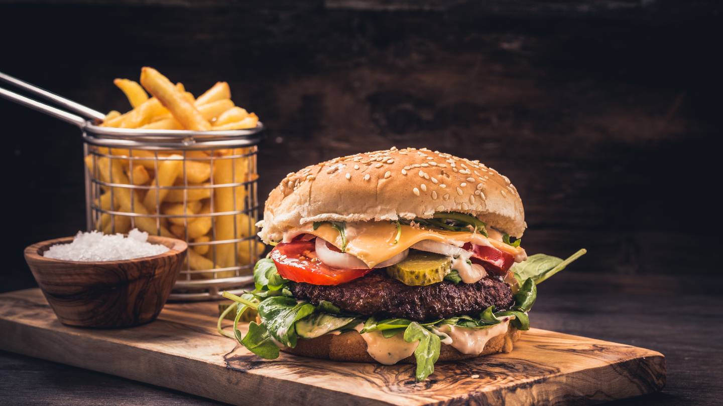 Cheeseburger with lettuce, tomato, pickles, onions, and sauce on a wooden board, with French fries in a small metal basket and a bowl of salt.