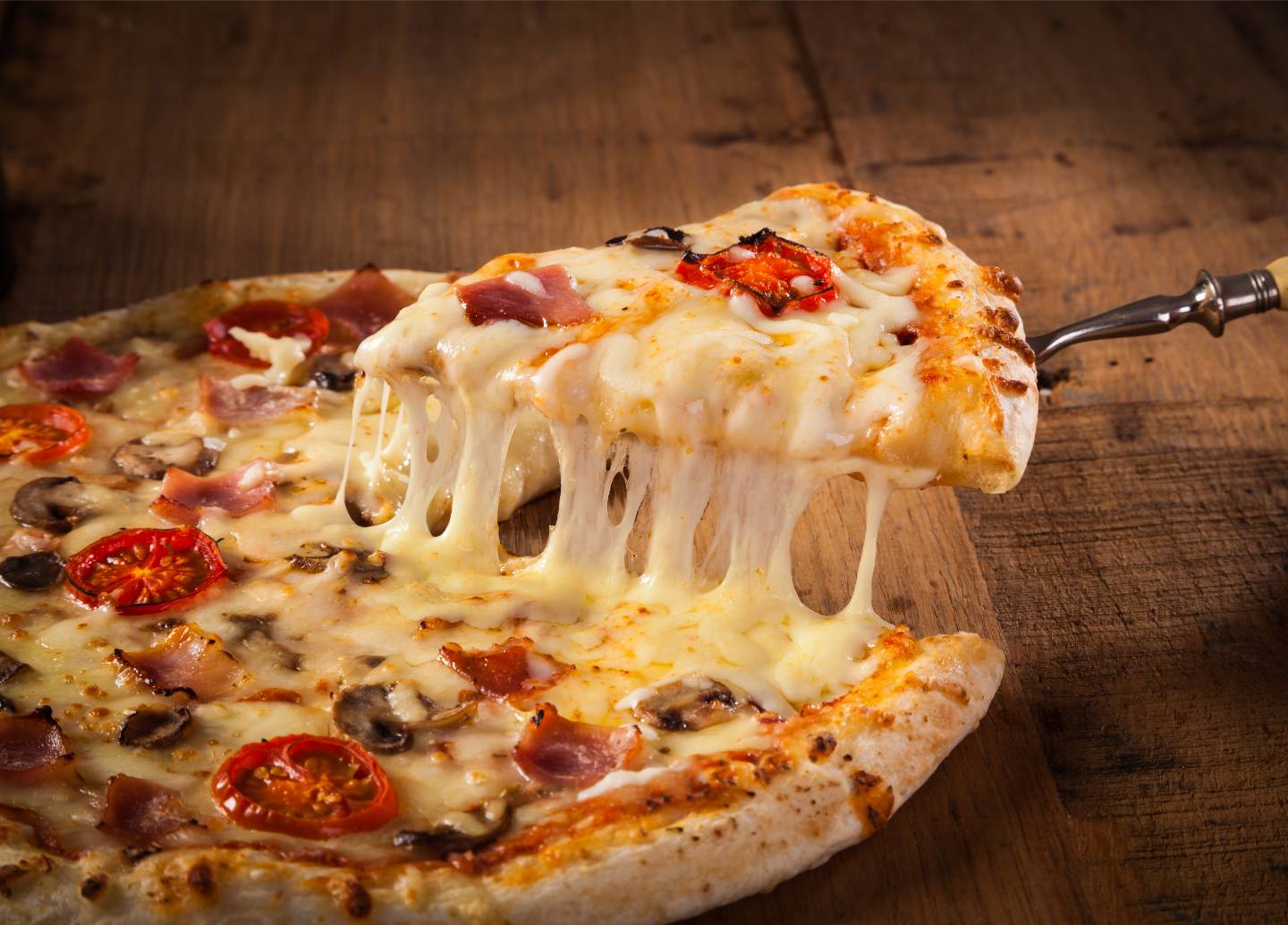 Slice of pizza with melted cheese, ham, mushrooms, and tomato being lifted from a whole pizza on a wooden surface.