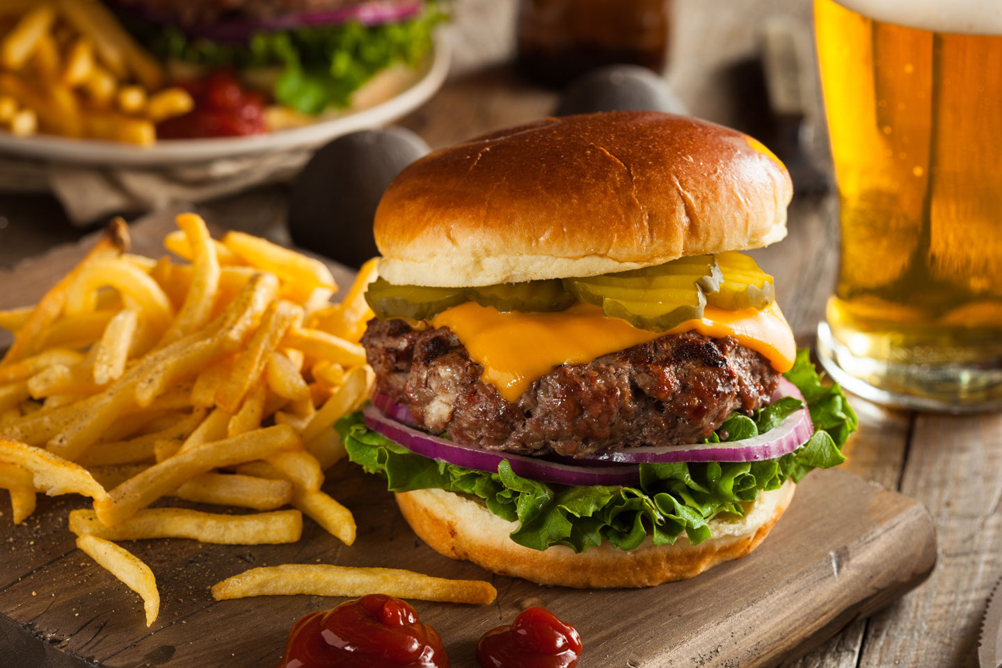 Cheeseburger with lettuce, red onion, pickles, and melted cheddar cheese on a bun served with seasoned French fries, ketchup, and a glass of beer.