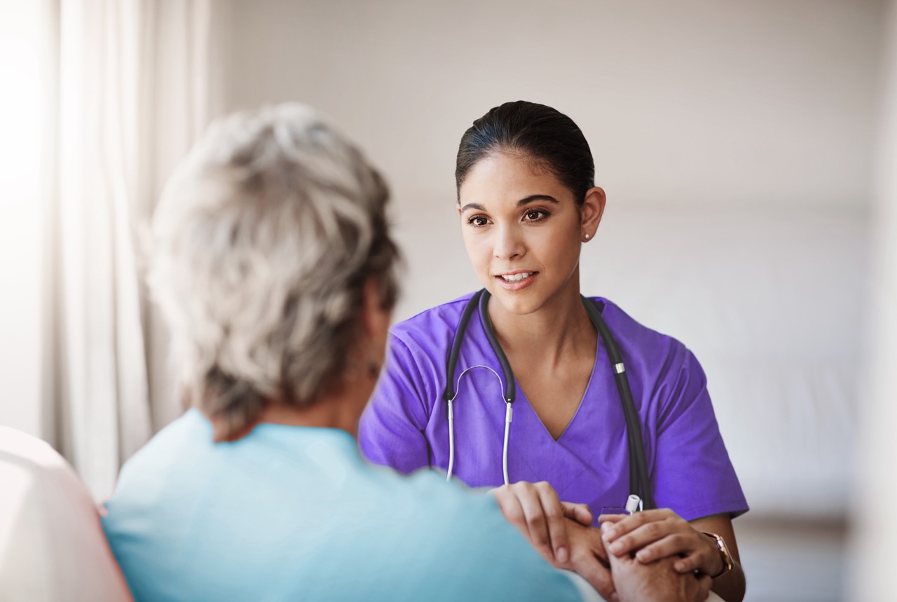 Female nurse in purple scrubs holding an elderly man’s hand while attentively listening to him.