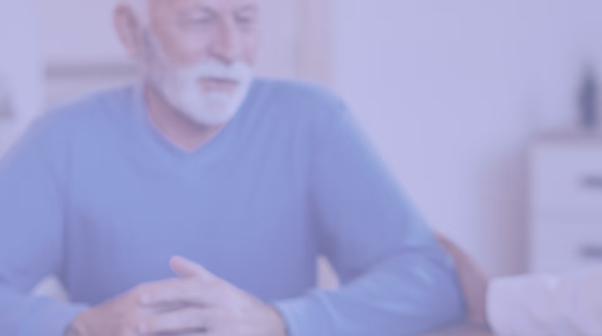 Blurred image of an older man with white beard and blue sweater sitting with clasped hands indoors.