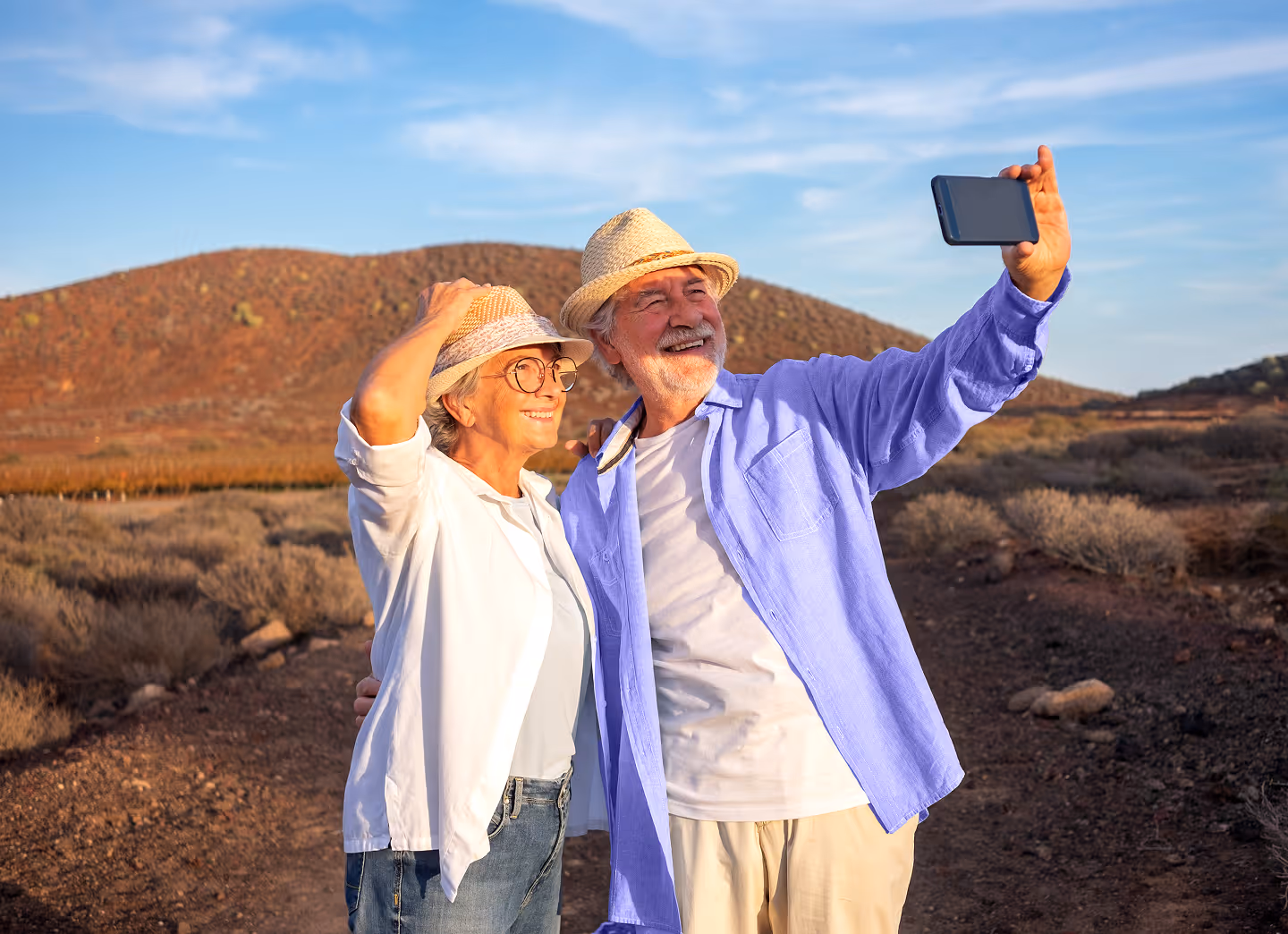 Smiling older couple in hats outdoors taking a selfie with a smartphone under blue sky.