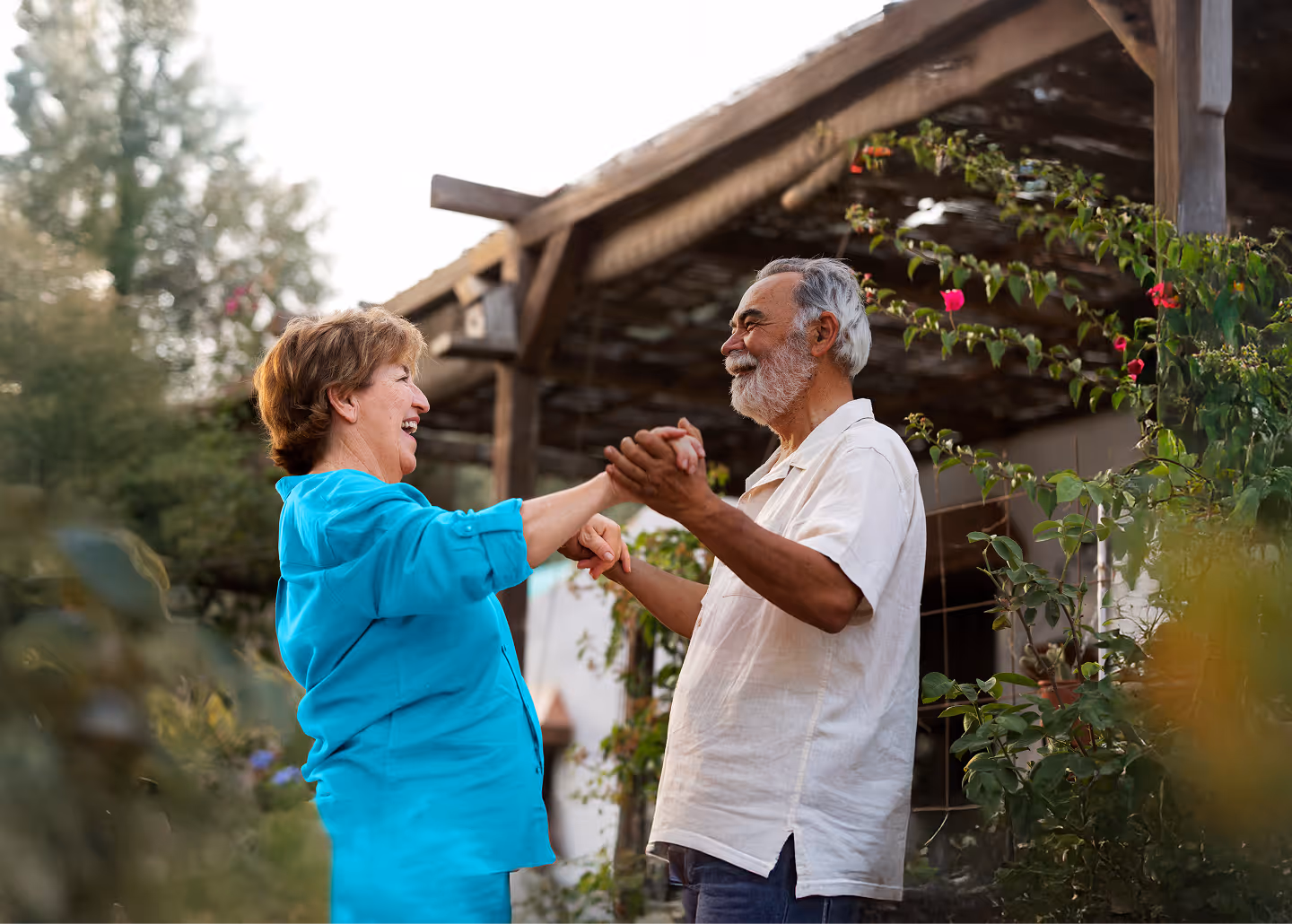 Elderly couple smiling and holding hands while dancing outdoors near a garden and wooden structure.