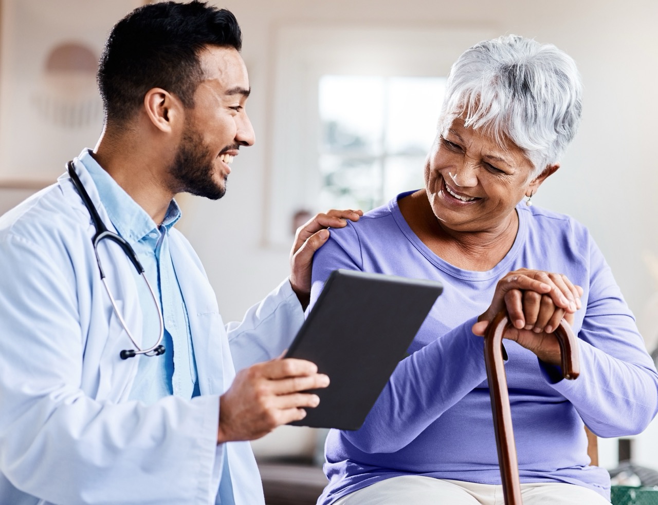 Smiling doctor holds a tablet and gently touches shoulder of happy elderly woman with a cane.