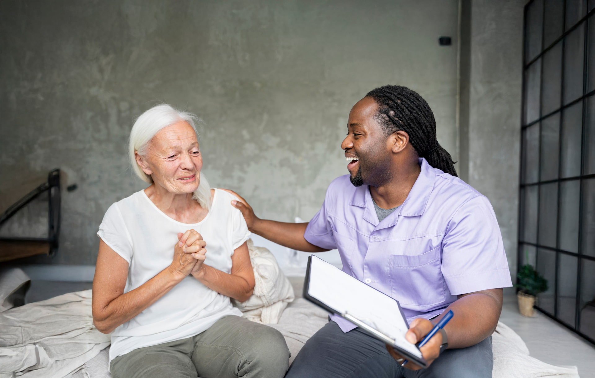 Caregiver in purple shirt smiling and holding clipboard while comforting elderly woman on bed.