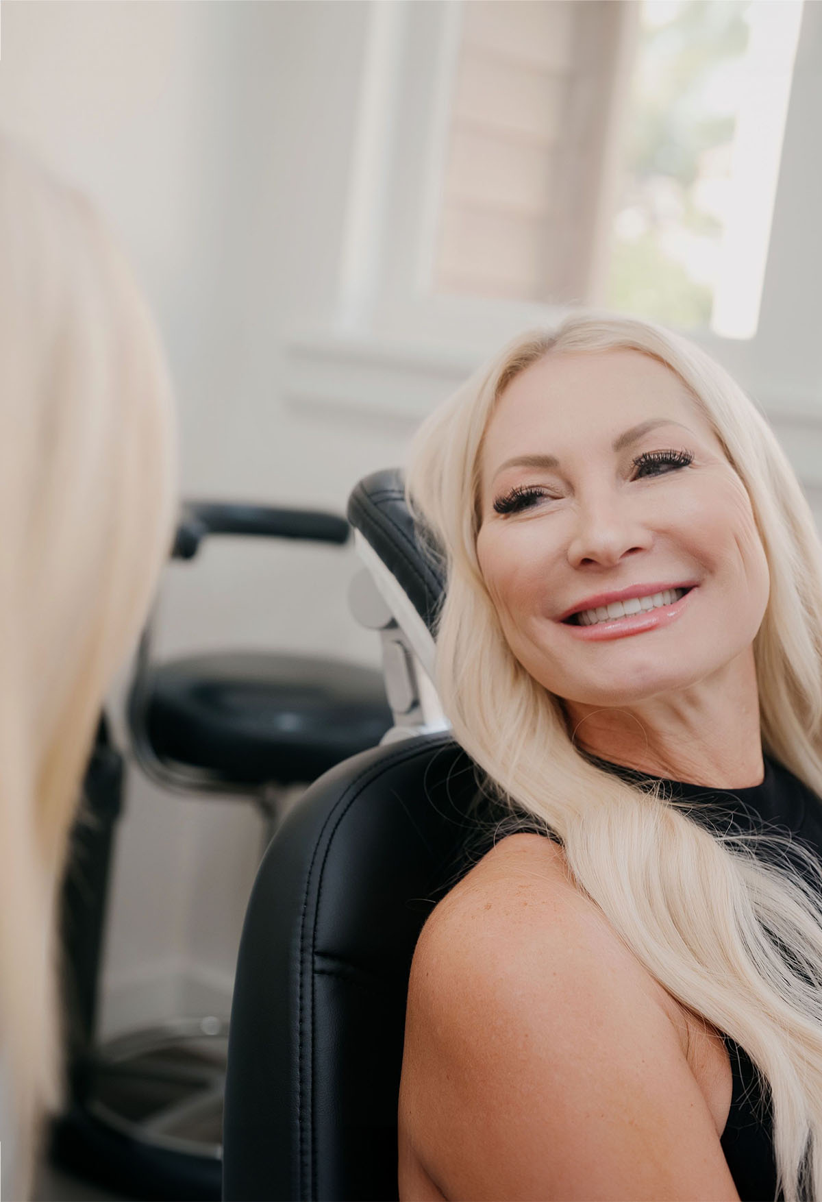 Smiling blonde woman with long hair sitting in a black chair, looking to the left.