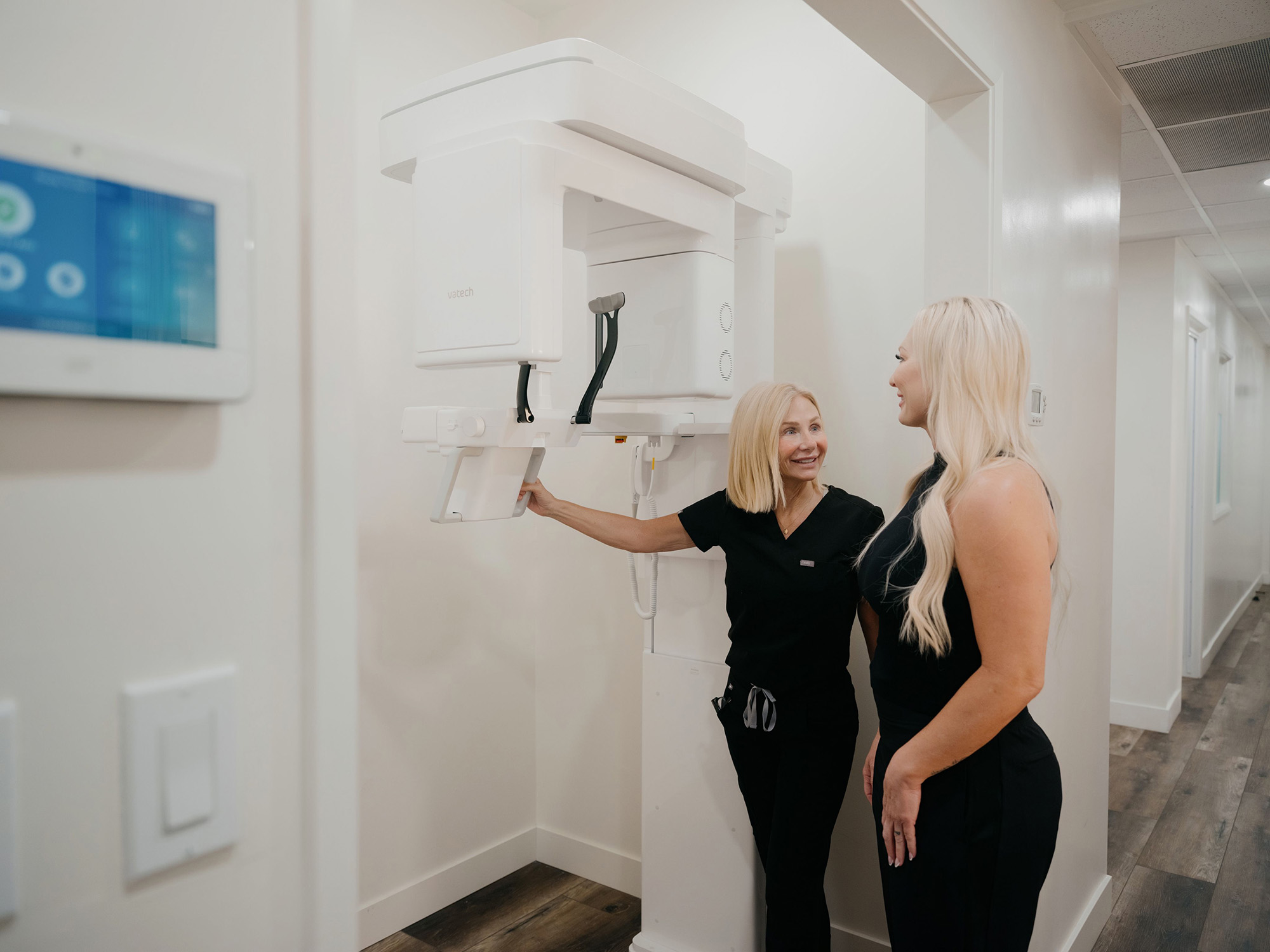 A medical professional shows a woman how to use a dental X-ray machine in a clinical hallway.