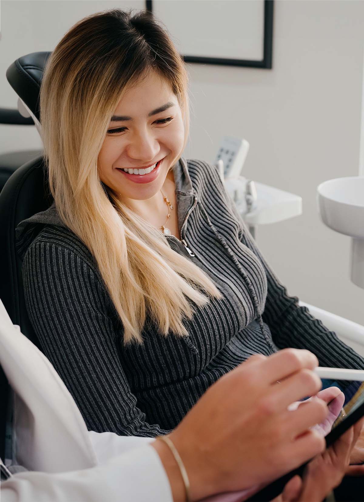 Smiling woman with blonde hair sitting in a chair looking at a tablet held by another person.