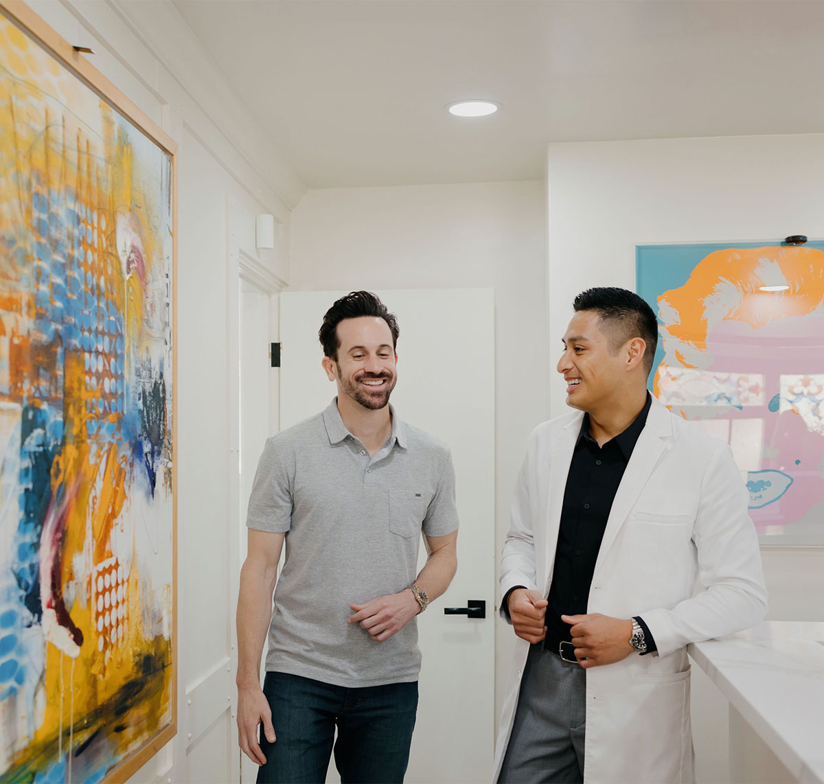 Man in gray polo shirt talking with man in white coat in a modern room with colorful artwork.