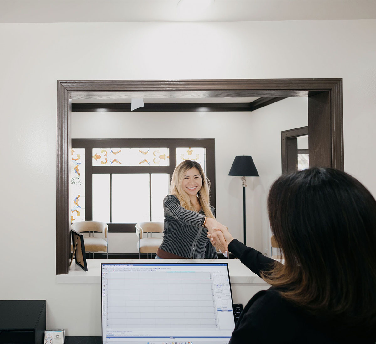 Two women shaking hands across a reception desk in a bright office with chairs and a lamp in the background.