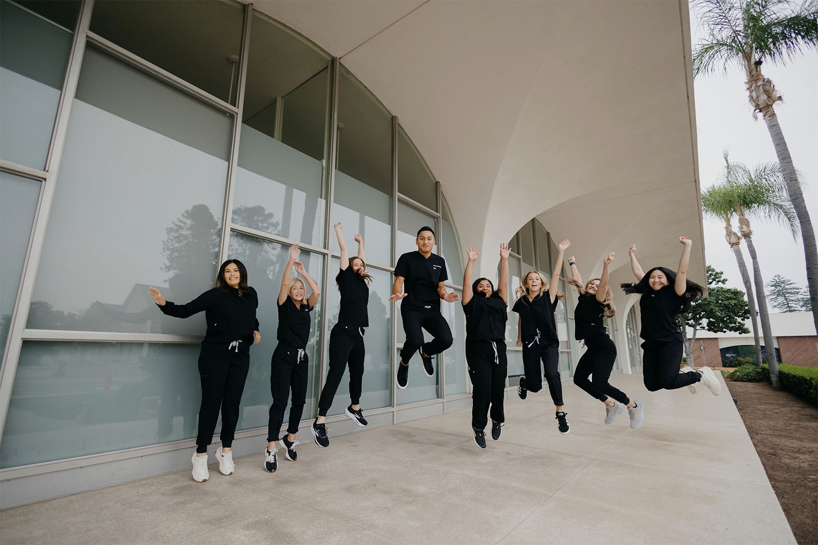 Seven women and one man in black scrubs jumping in unison outside a modern building with glass windows.