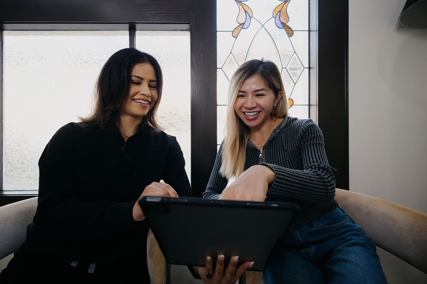 Two women sitting and smiling while looking at a digital tablet together indoors.