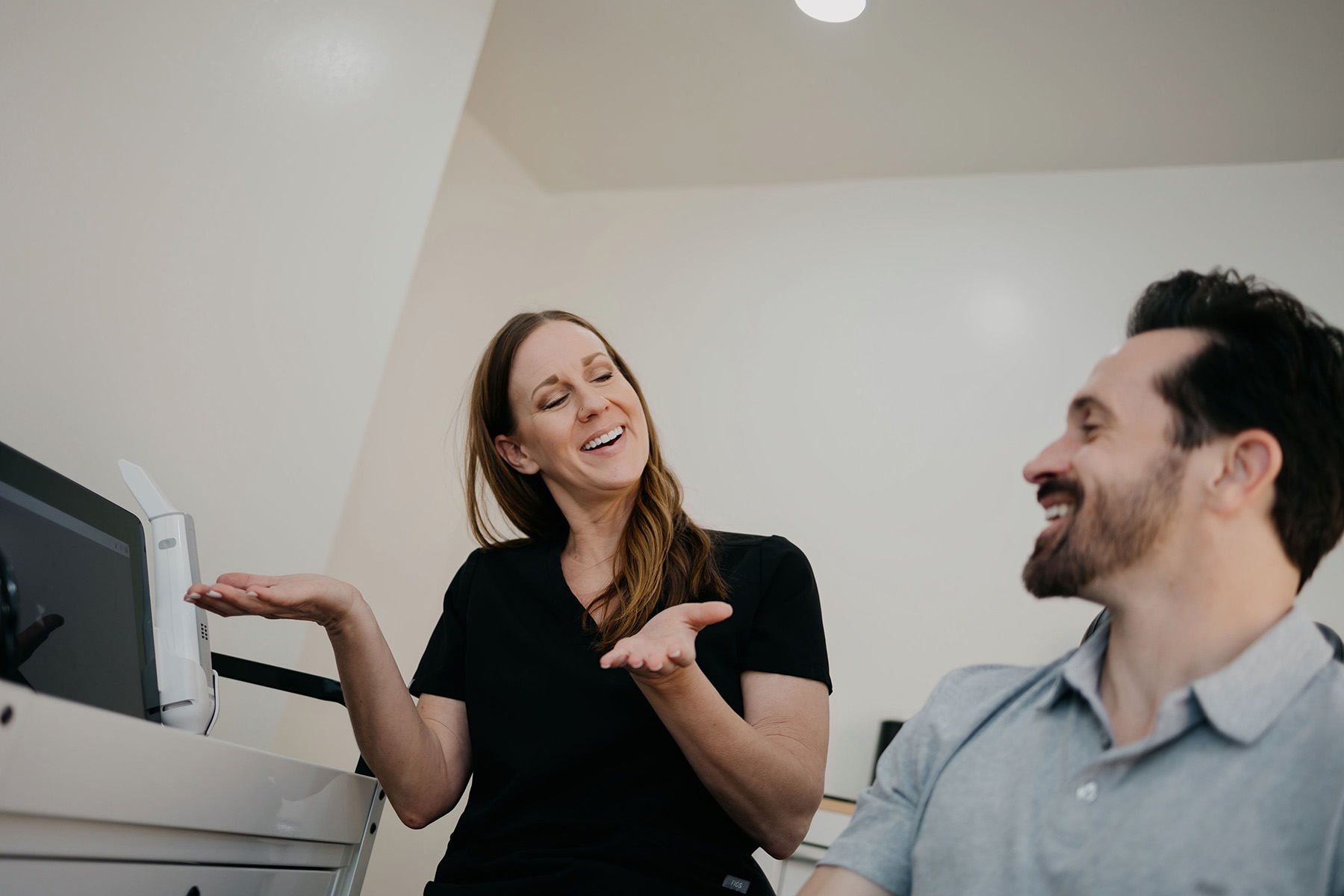 Smiling woman in black scrubs talking and gesturing to a smiling man in a light gray shirt.