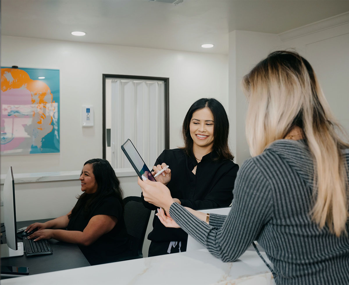Two women at a reception desk helping a blonde-haired woman with a tablet device in a modern office.