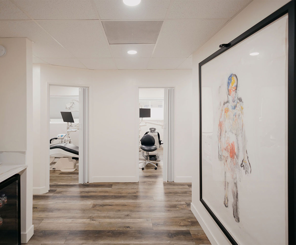 Hallway with wooden floor, two open rooms with dental chairs, and a large framed abstract painting on the wall.