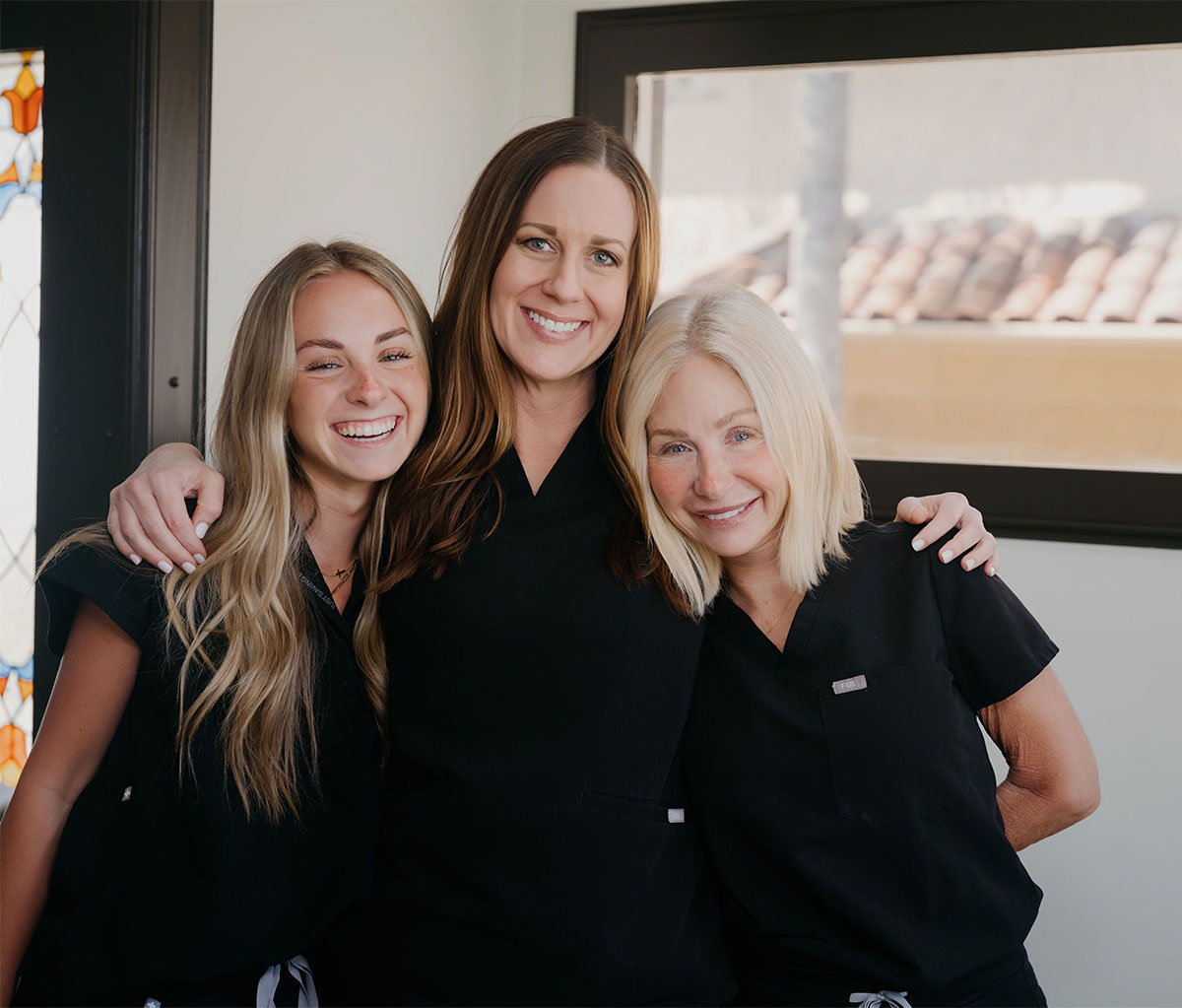 Three smiling women in black scrubs standing close together indoors near a window and stained glass door.