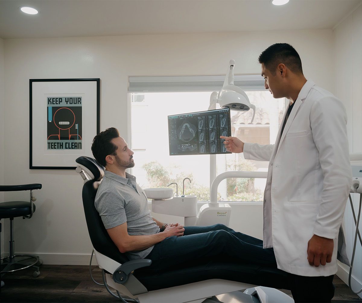 Dentist in white coat showing dental X-rays on a screen to a seated male patient in a dental office.