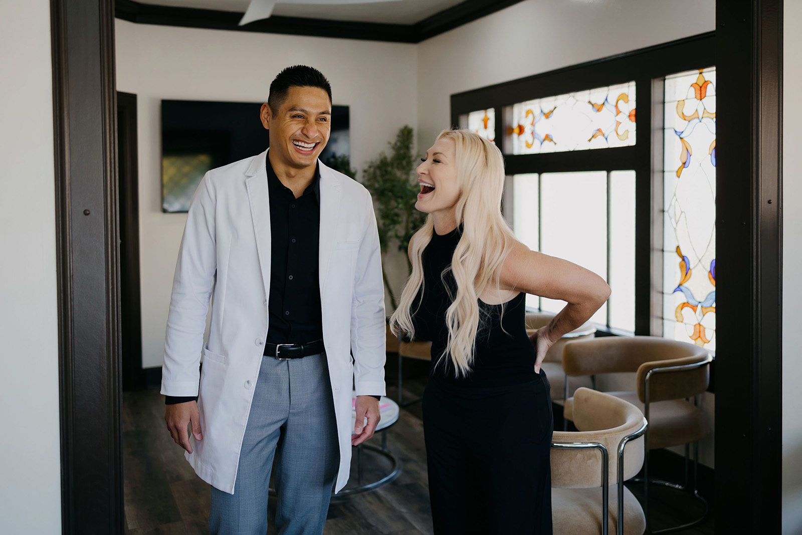 Man in a white coat and woman in black laughing together in a room with stained glass windows.