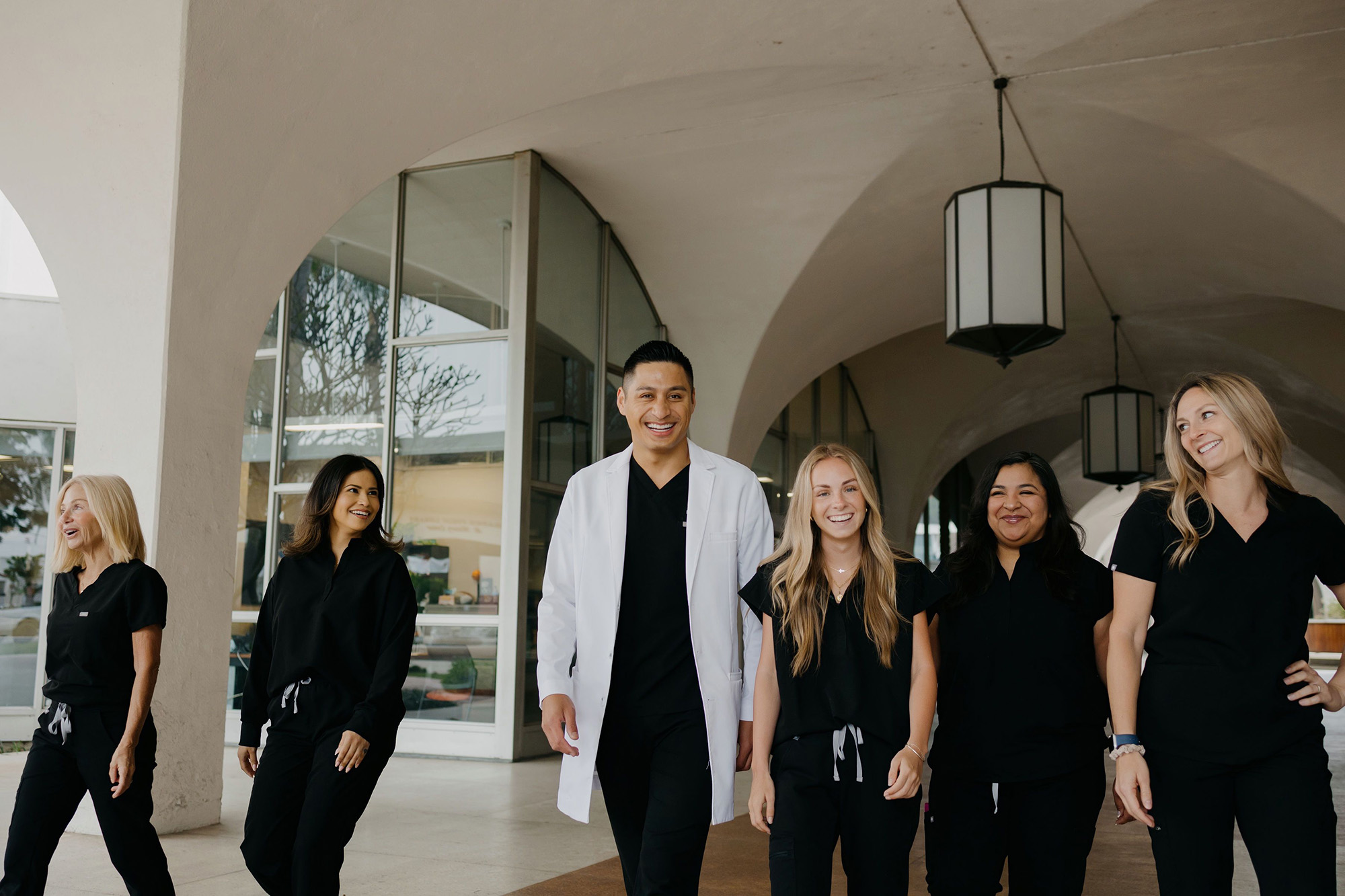Five medical professionals walking and smiling under an arched walkway, wearing black scrubs and one white coat.