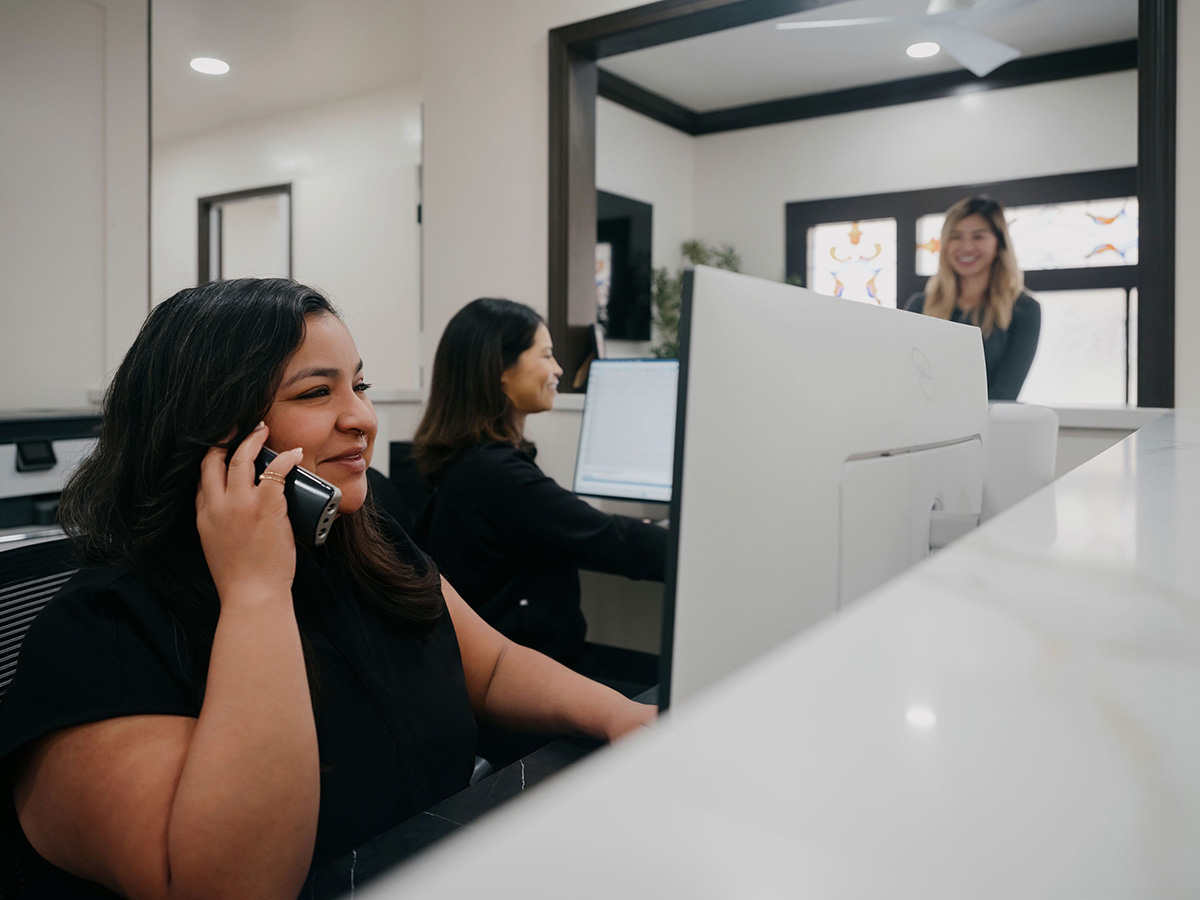 Two women at desks with computers; one talks on phone, another stands behind a counter smiling.