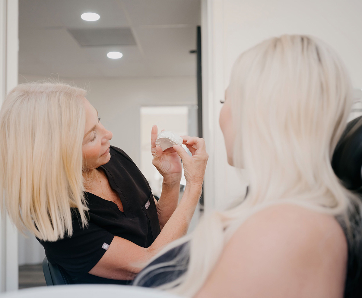 Blonde dental professional shows a dental mold to a seated blonde patient in a clinic.
