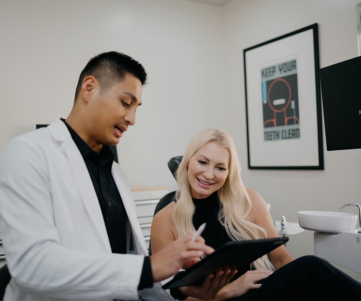 Dentist in white coat showing dental information on a tablet to a smiling female patient.