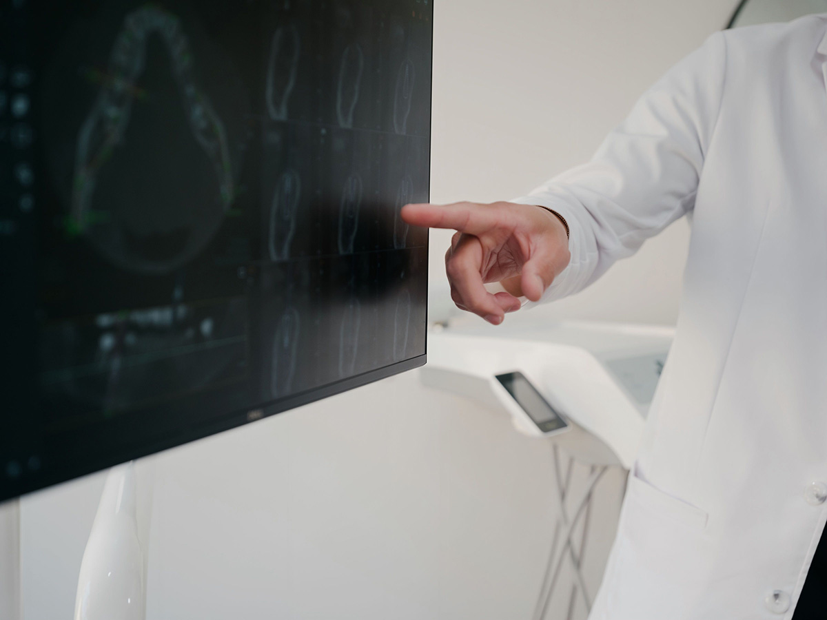 Person in white coat pointing at medical images on a dark monitor screen in a clinical setting.