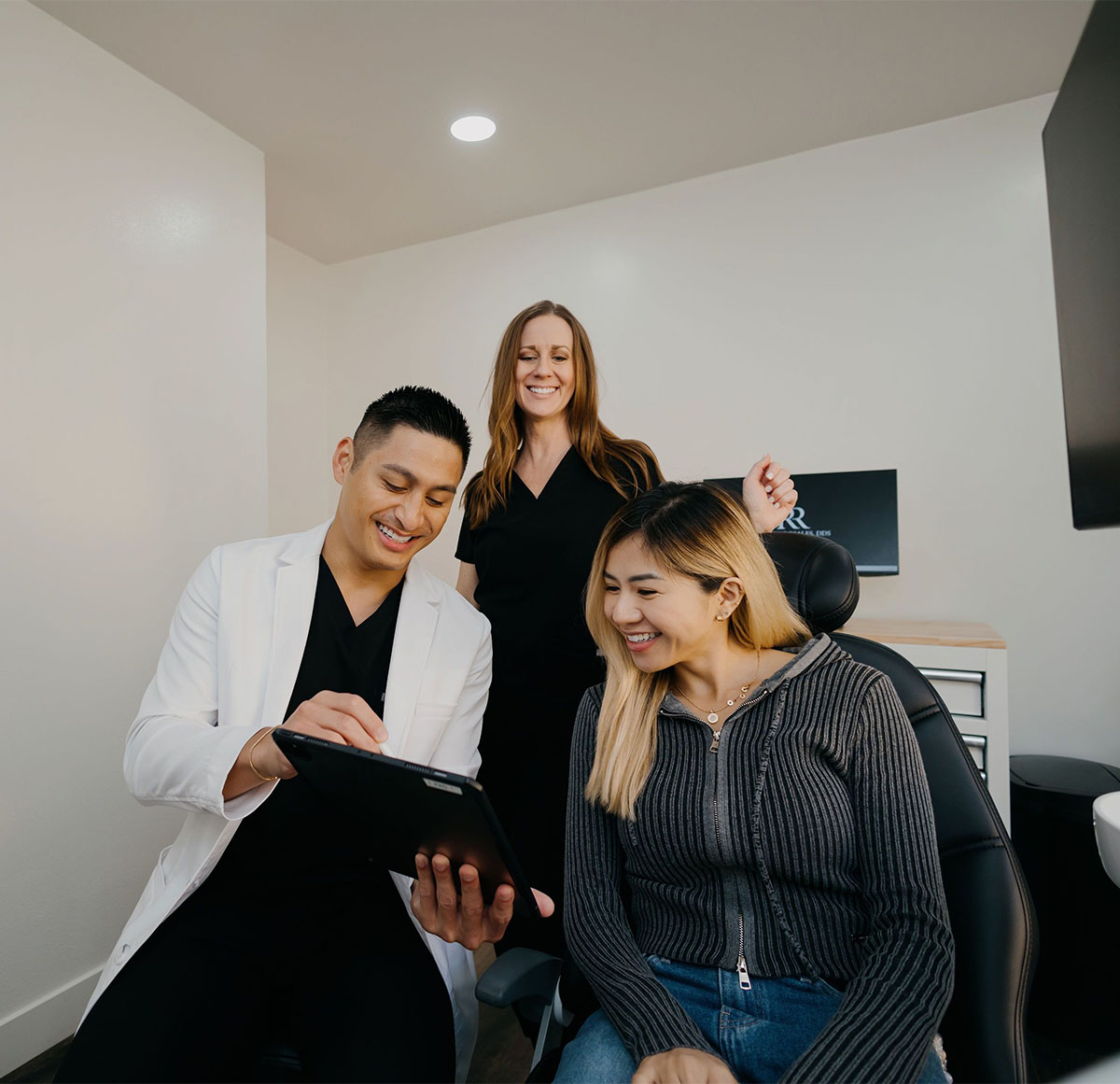 Dentist and assistant smiling while showing a seated female patient information on a tablet.