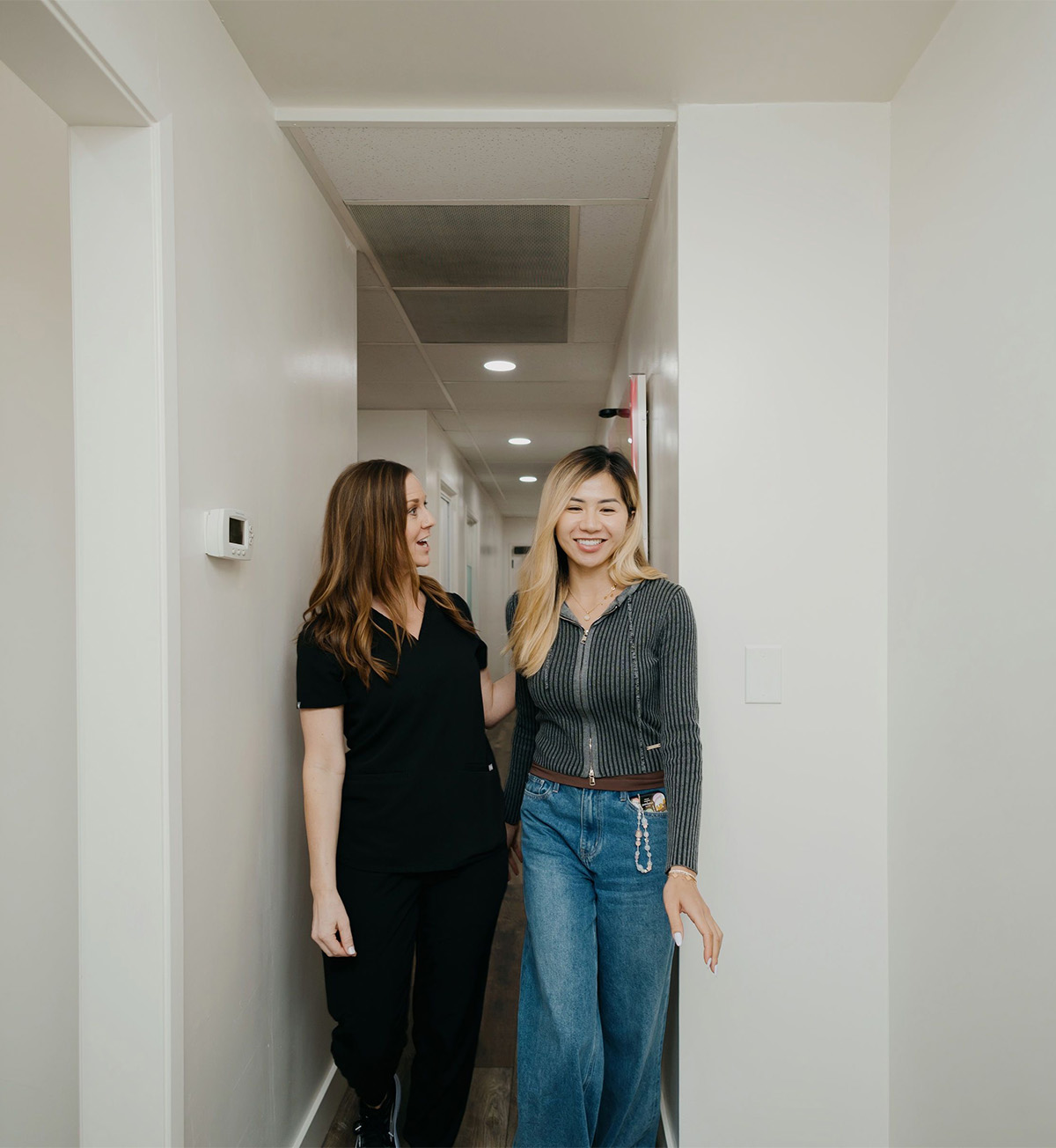 Woman in black scrubs walks and talks with another woman in jeans and a gray sweater in a hallway.