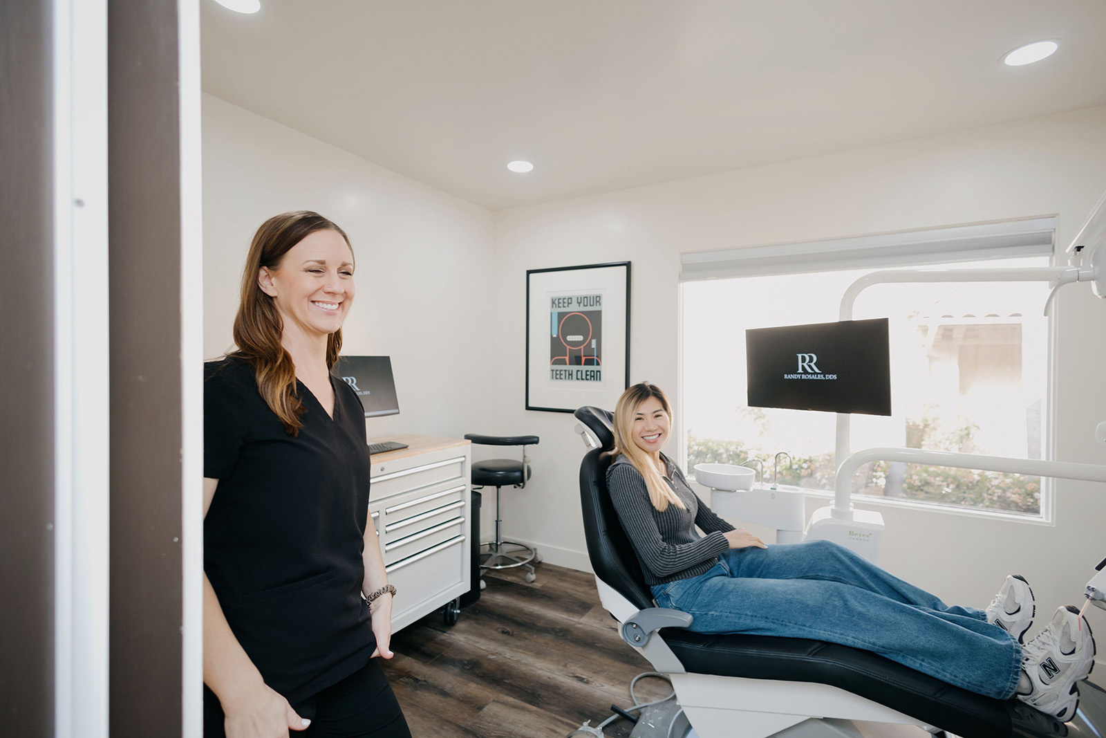 Smiling dental patient reclined in chair and a staff member standing smiling in a bright dental office.