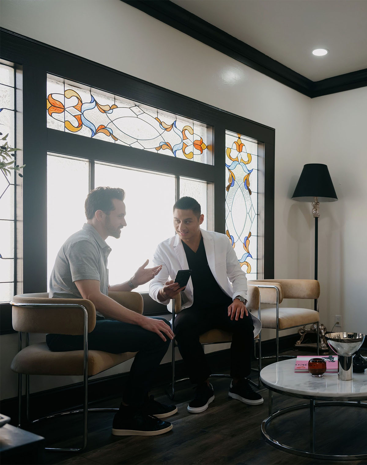 Two men seated in a modern room with stained glass windows, one showing a phone to the other.
