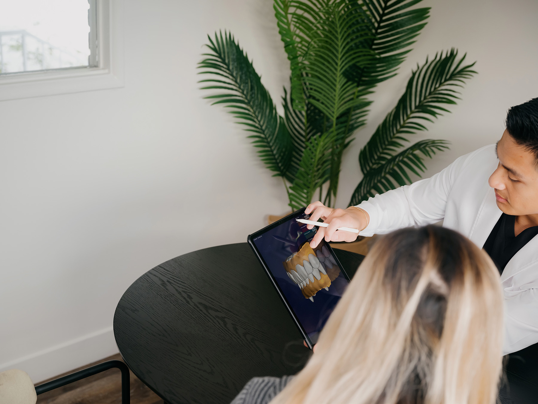 Dentist shows a dental 3D model on a tablet to a patient at a black round table.