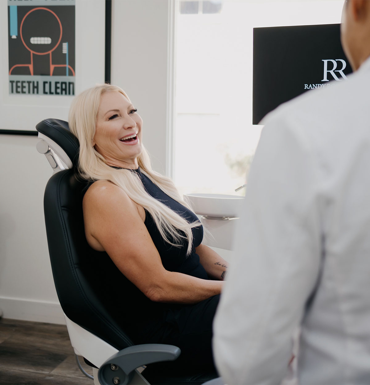 Smiling woman with long blonde hair sitting in a dental chair talking to a person in white coat.
