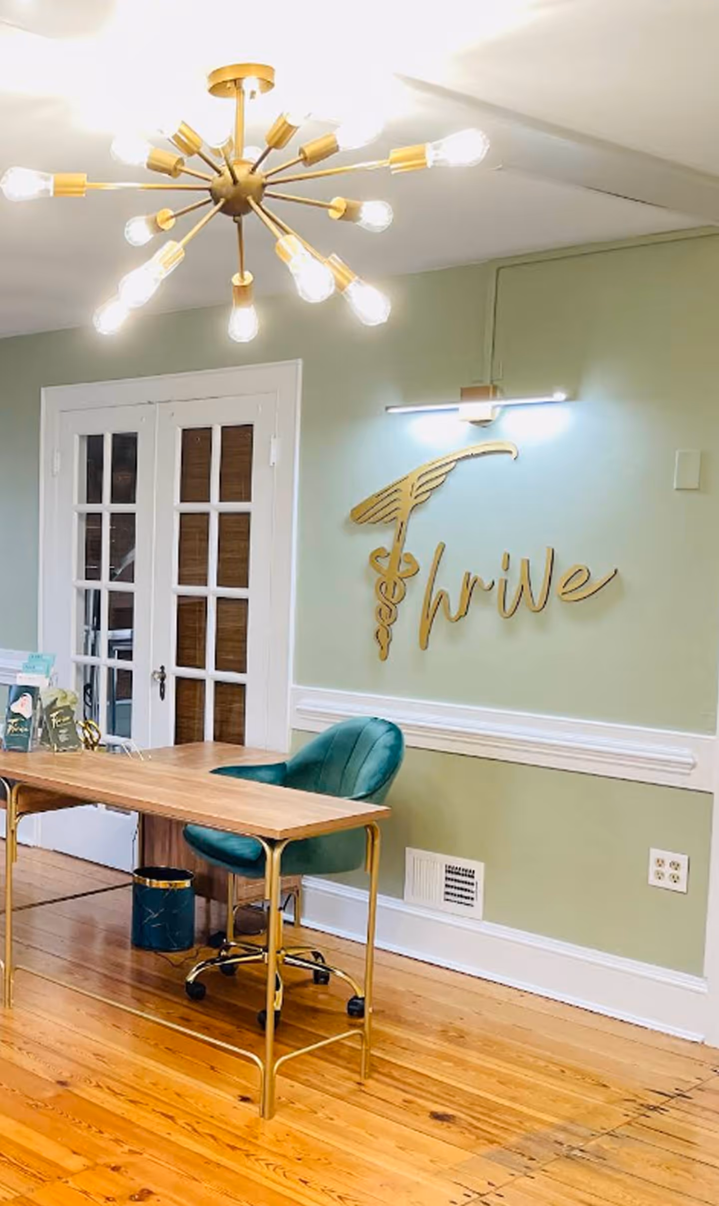 Office with gold chandelier, wooden desk, green chair, and gold Thrive sign on green wall.