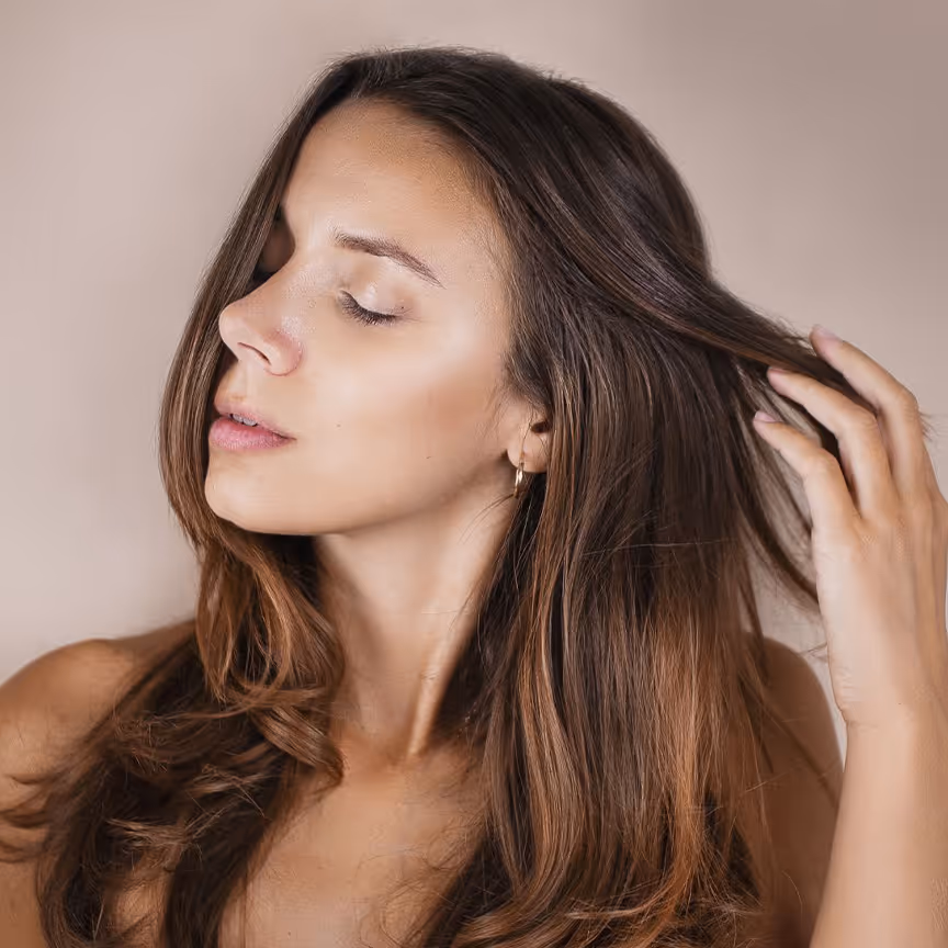 Woman with long brown hair touching it gently with eyes closed, facing left on beige background.