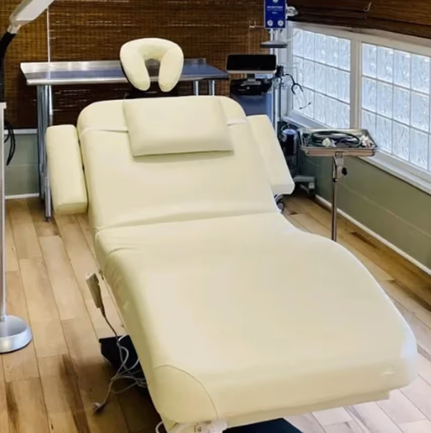 Cream-colored adjustable medical or massage table in a well-lit room with wooden floor and equipment nearby.