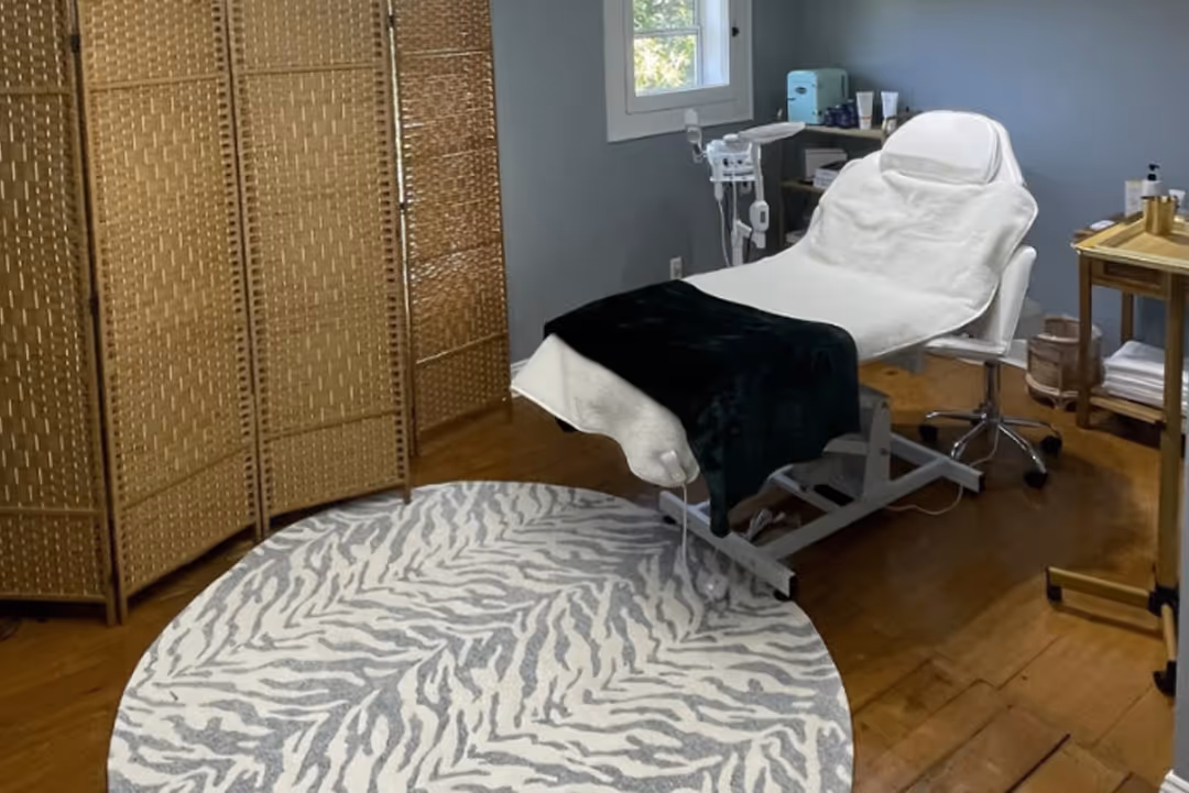 Treatment room with white spa bed, black blanket, round patterned rug, and wooden screen divider.