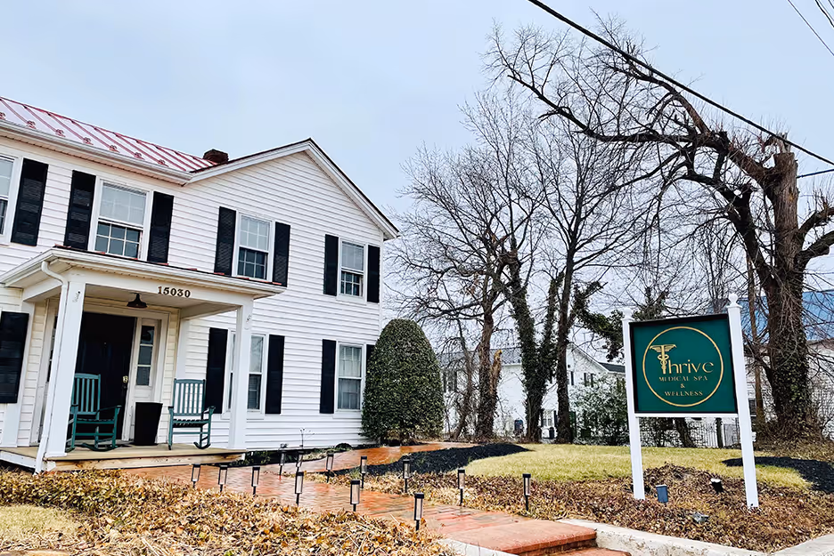 White two-story house with black shutters and Thrive Medical Spa sign on a lawn with leafless trees.