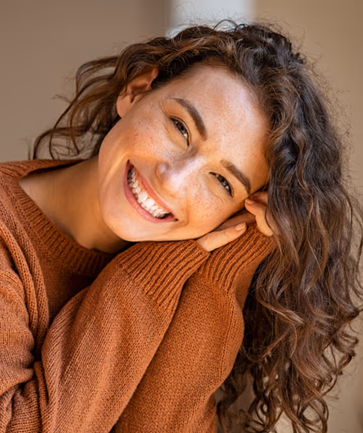 Smiling woman with curly hair leaning her head on her hands wearing a brown knit sweater.